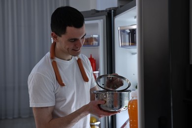 Photo of Man with sausages and pot near refrigerator in kitchen at night