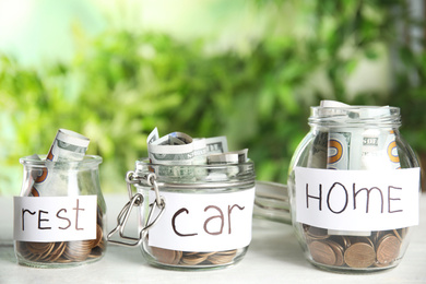 Photo of Glass jars with money and tags REST, CAR, HOME on white table against blurred background