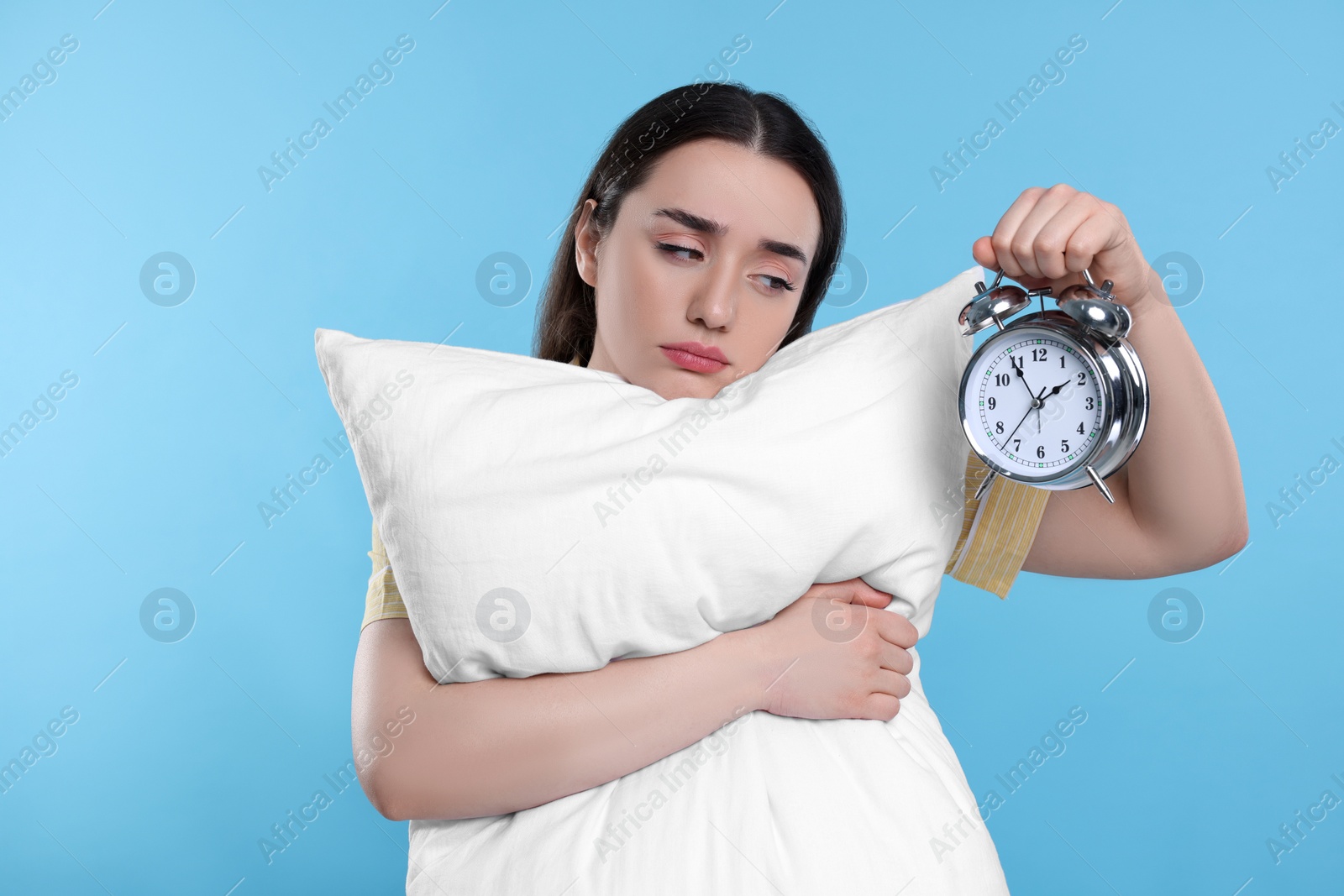 Photo of Tired young woman with pillow and alarm clock on light blue background. Insomnia problem