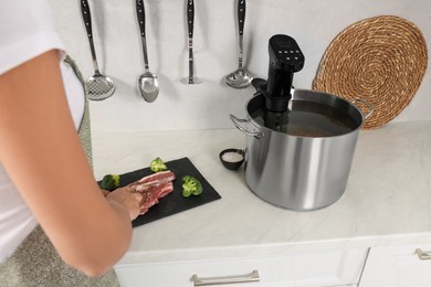 Photo of Woman cutting meat near pot with sous vide cooker in kitchen, closeup. Thermal immersion circulator