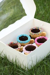 Photo of Box of delicious edible biscuit coffee cups decorated with sprinkles on green grass outdoors