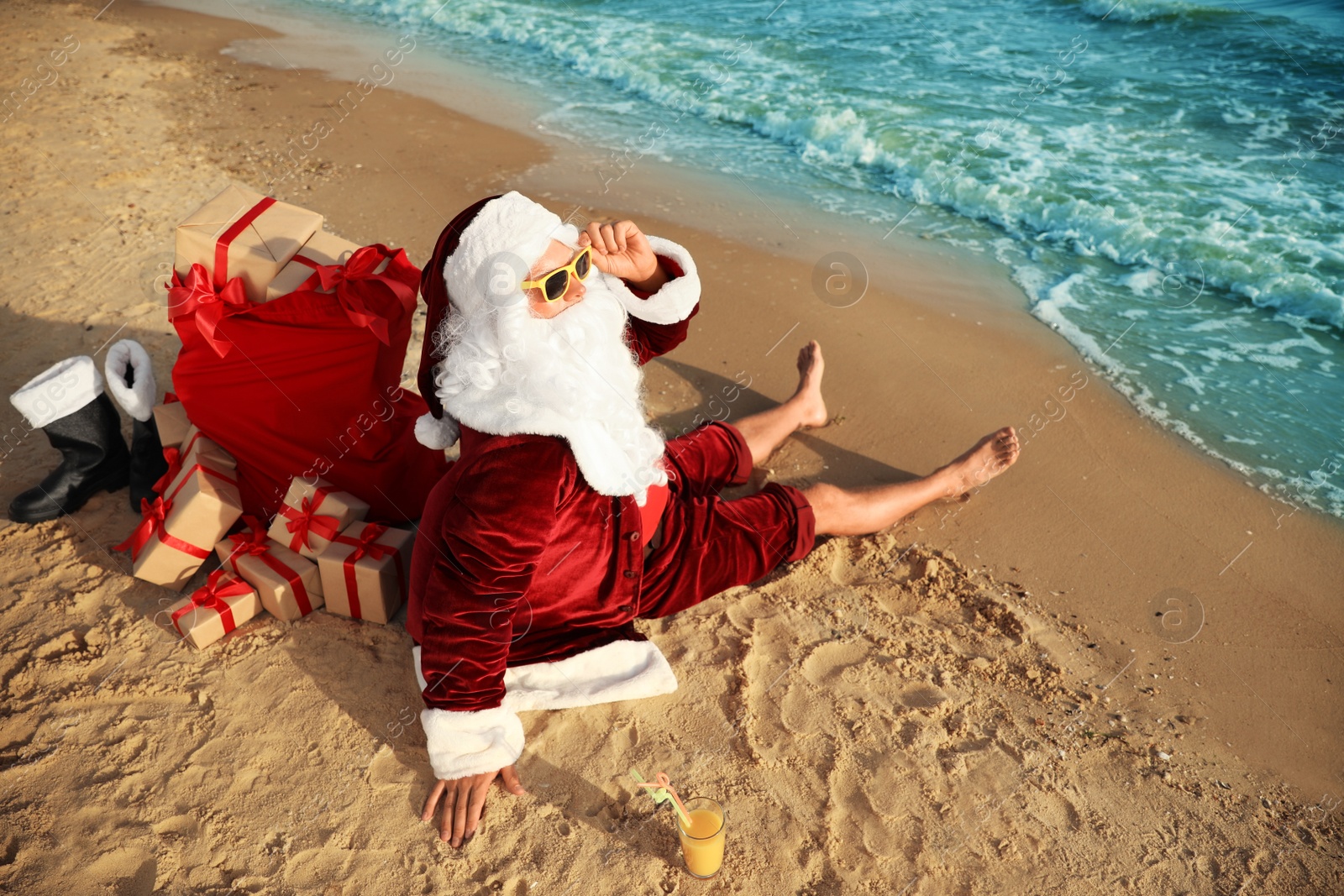 Photo of Santa Claus with cocktail and bag of presents relaxing on beach. Christmas vacation