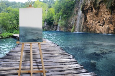 Wooden easel with picture on bridge over lake and beautiful view of waterfall 