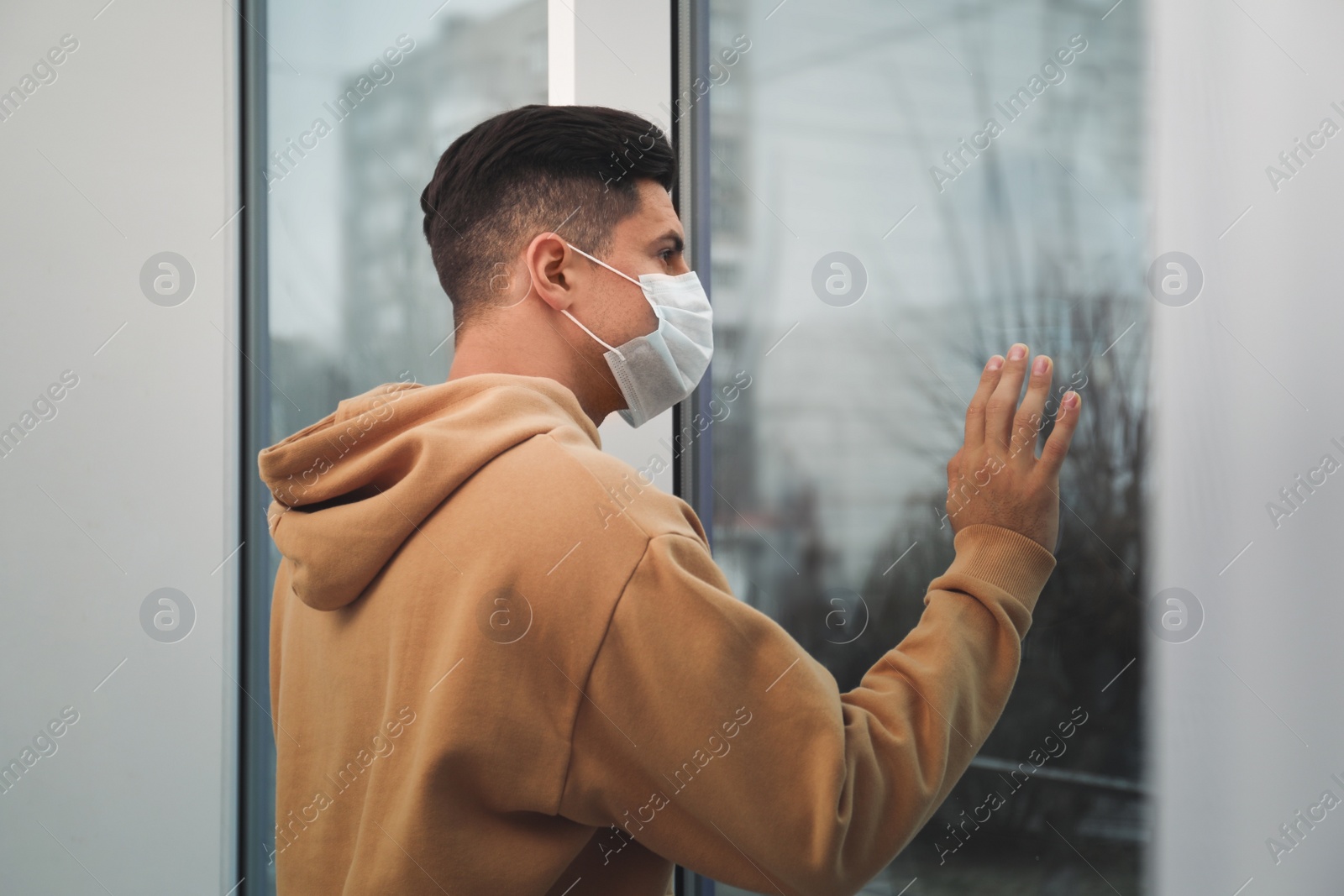 Photo of Sad man in protective mask near window indoors. Self-isolation during coronavirus pandemic