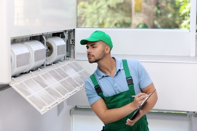 Photo of Professional technician with clipboard maintaining modern air conditioner indoors