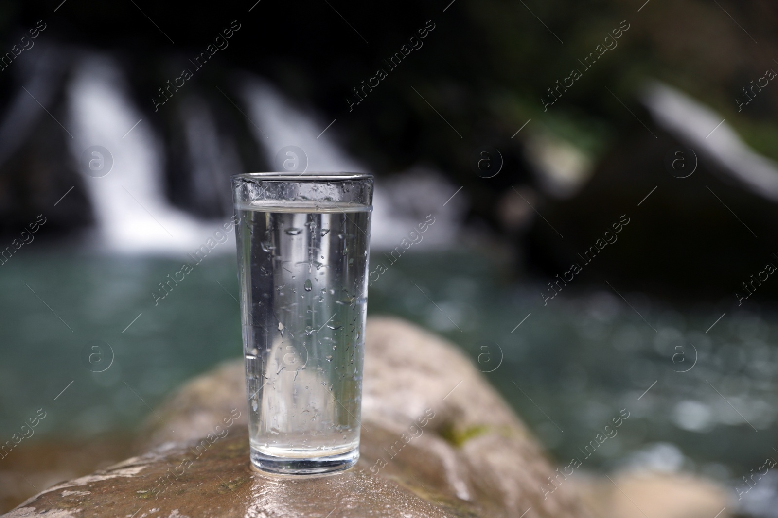 Photo of Glass of water on stone near waterfall outdoors. Space for text