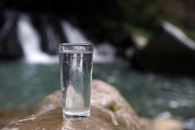 Glass of water on stone near waterfall outdoors. Space for text