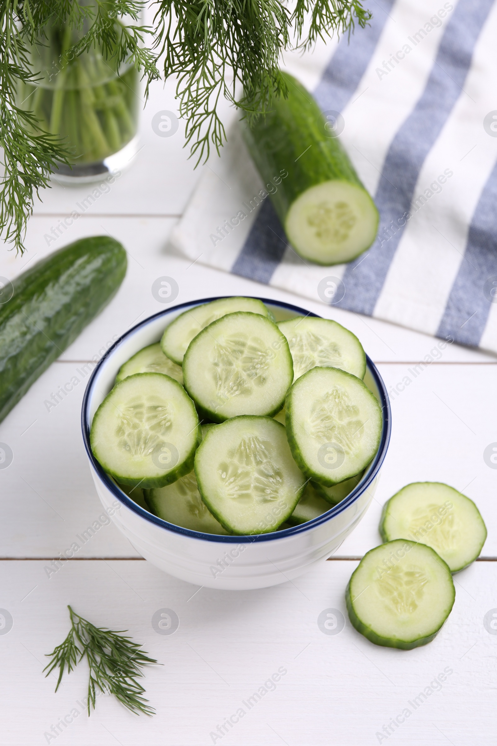 Photo of Cut cucumber in bowl, fresh vegetables and dill on white wooden table, above view