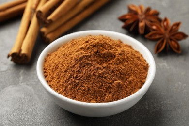 Photo of Bowl of cinnamon powder, sticks and star anise on grey table, closeup