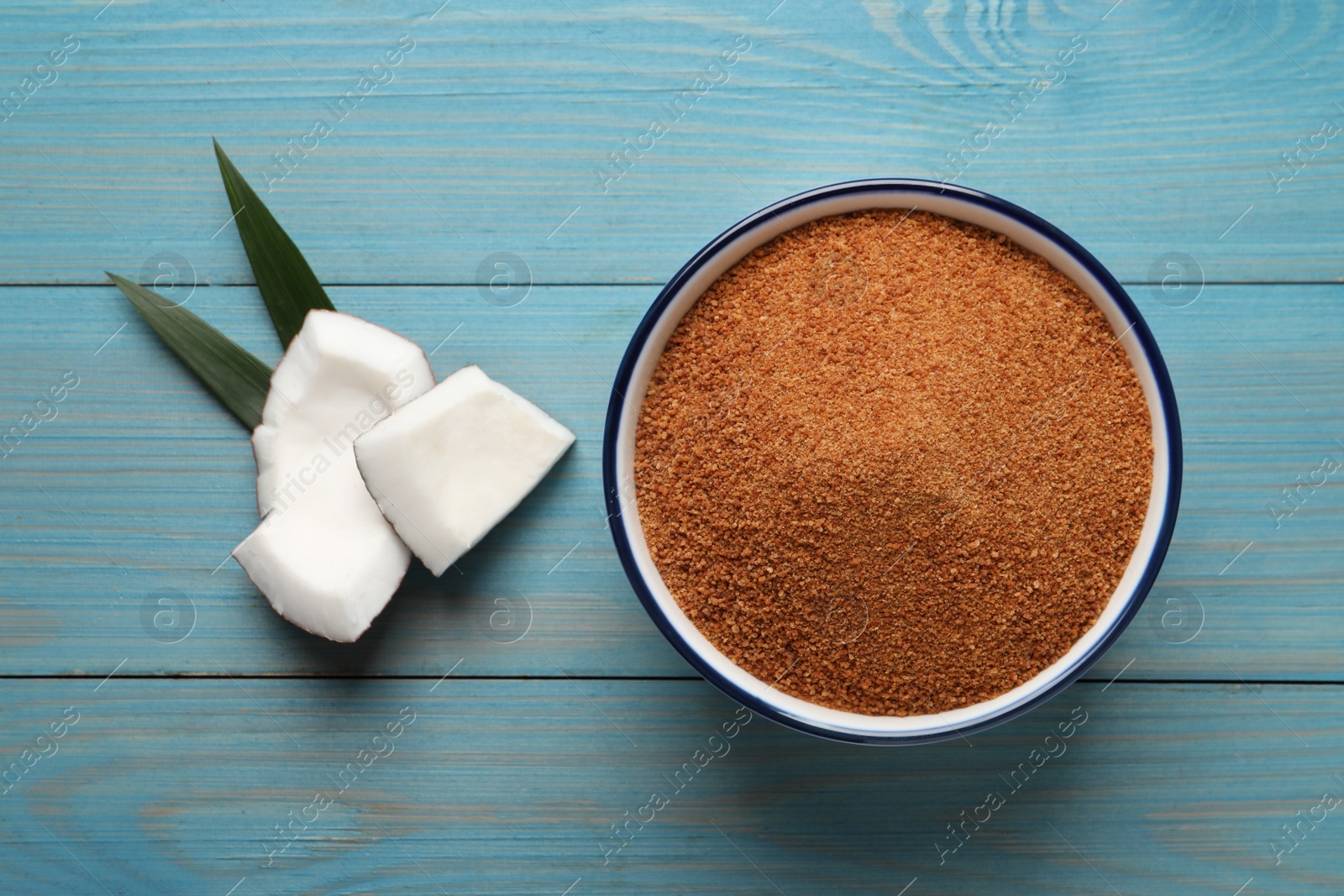 Photo of Natural coconut sugar in bowl on light blue wooden table, flat lay