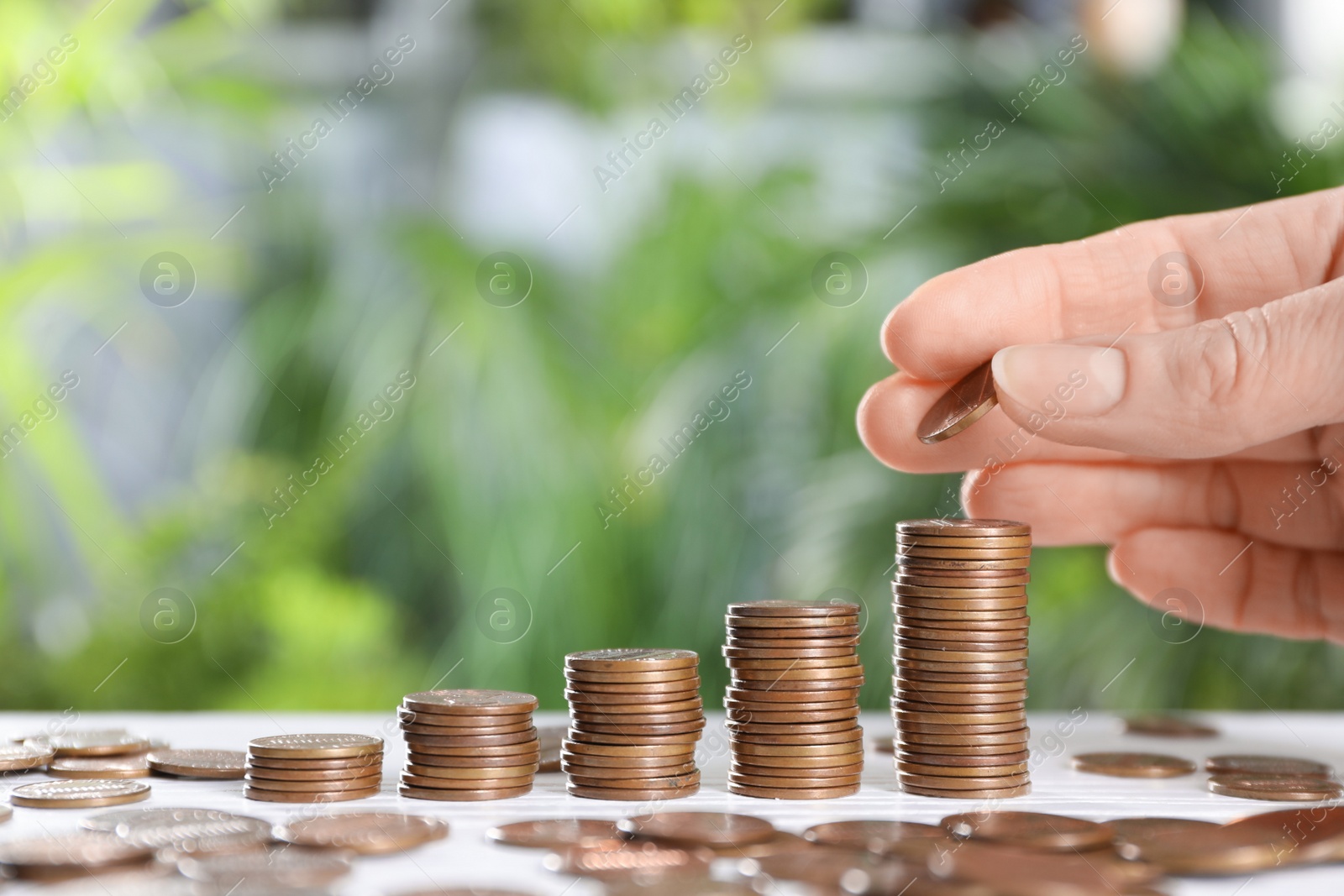 Photo of Woman stacking coins at white table against blurred green background, closeup. Space for text