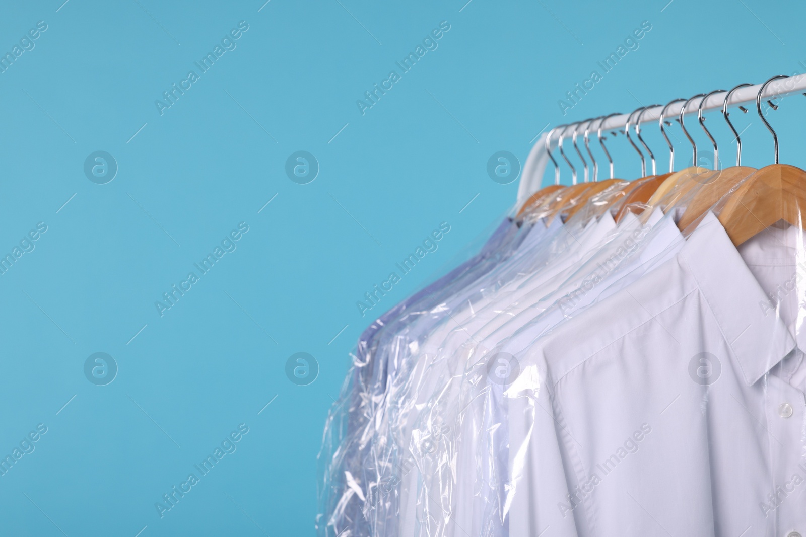 Photo of Dry-cleaning service. Many different clothes in plastic bags hanging on rack against light blue background, closeup and space for text