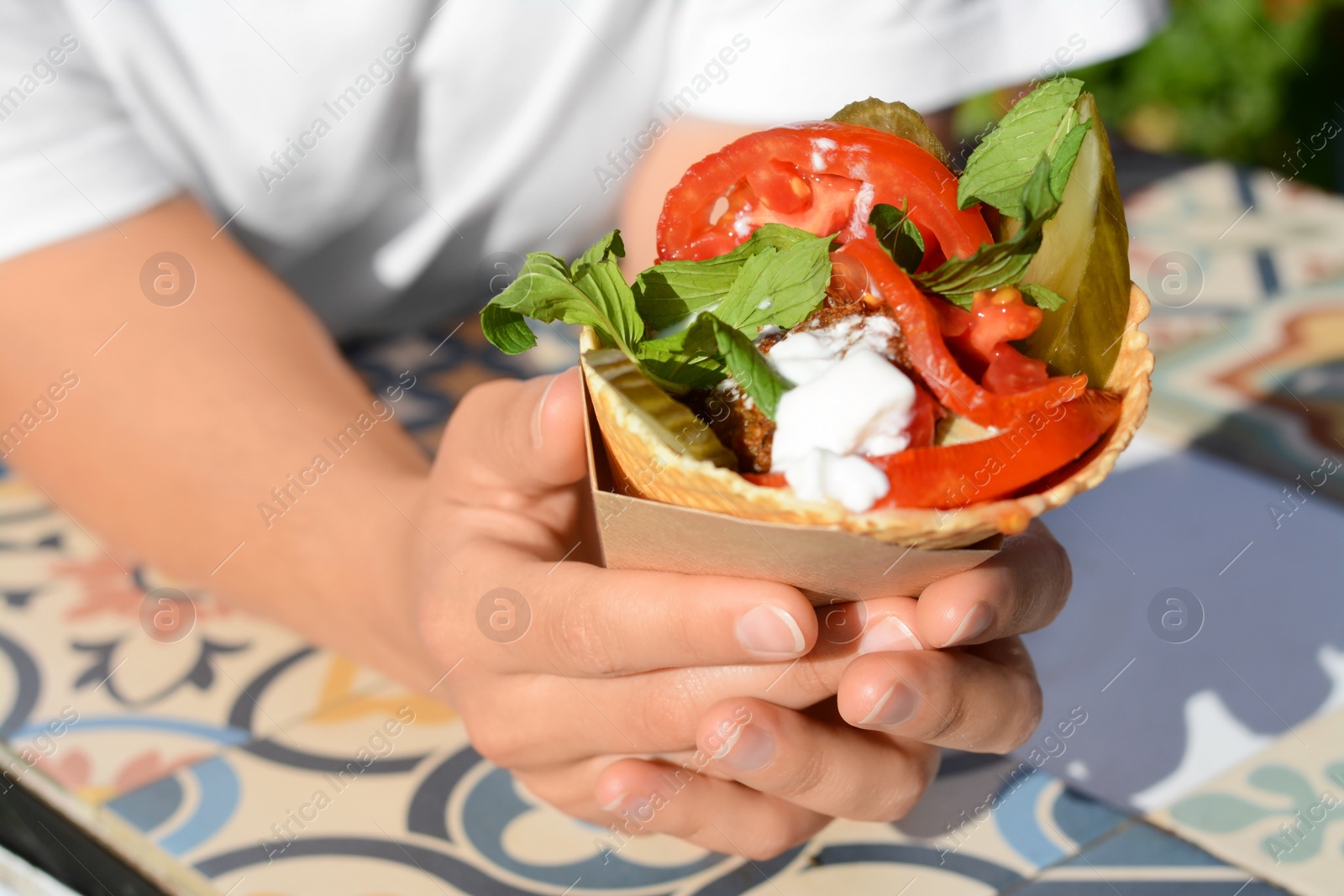 Photo of Woman holding wafer with falafel and vegetables at colorful tiled table outdoors, closeup. Street food