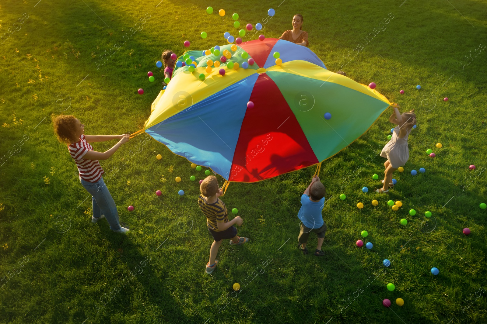 Image of Group of children and teachers playing with rainbow playground parachute on green grass, above view. Summer camp activity