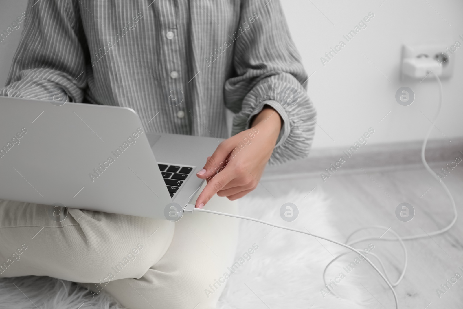 Photo of Woman connecting charger cable to laptop on floor near white wall, closeup