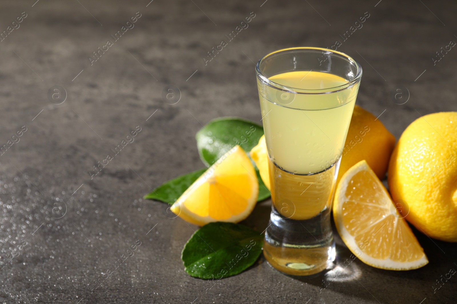 Photo of Tasty limoncello liqueur, lemons and green leaves on grey table, closeup. Space for text