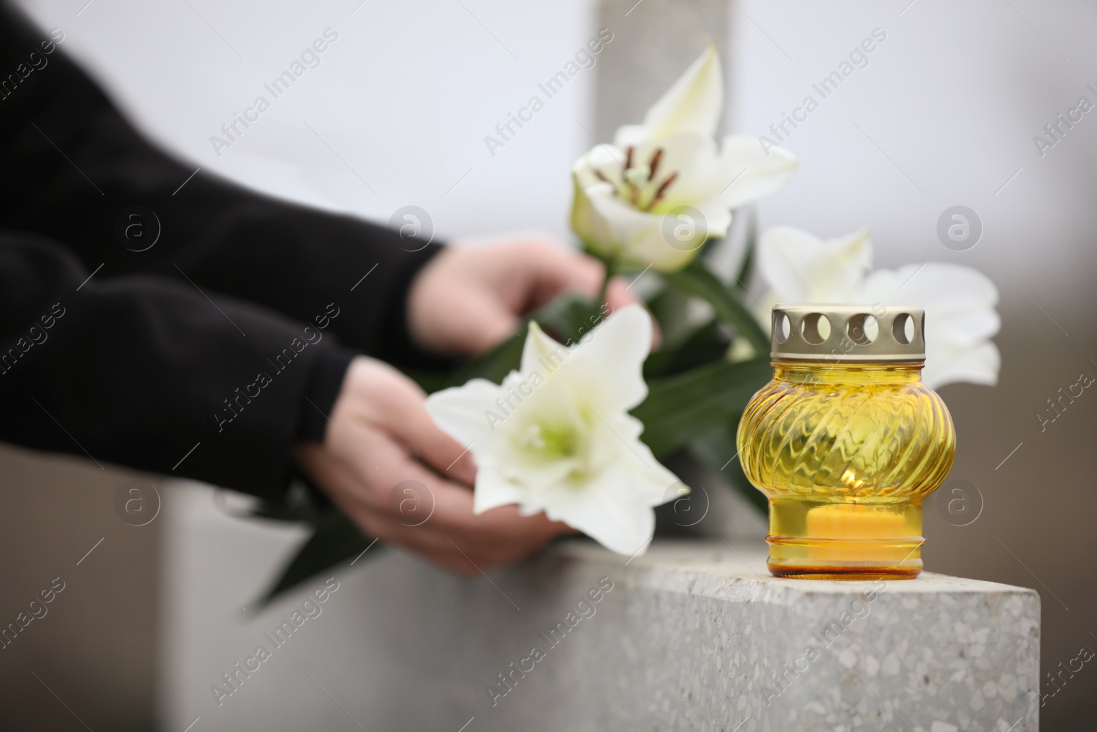 Photo of Woman holding white lilies near light grey granite tombstone with candle outdoors, closeup. Funeral ceremony