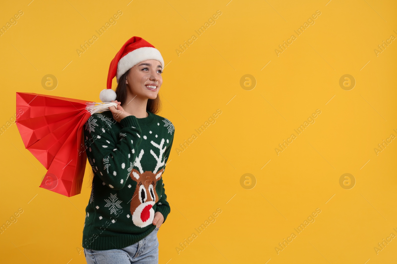 Photo of Happy young woman in Christmas sweater and Santa hat with shopping bags on orange background. Space for text