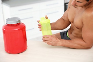 Photo of Young shirtless athletic man preparing protein shake at wooden table indoors, closeup view