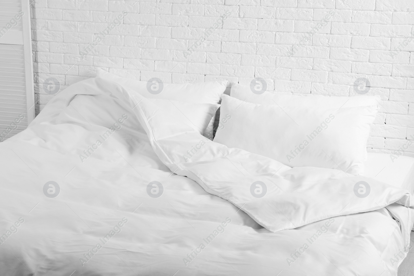 Photo of Large comfortable bed with pillows and blanket near white brick wall indoors. Stylish interior