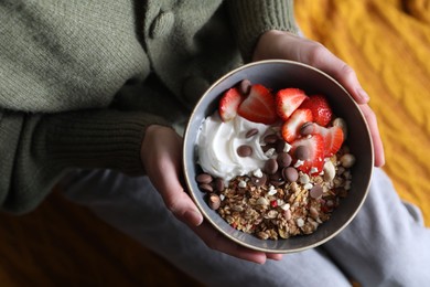 Photo of Woman holding bowl of tasty granola with chocolate chips, strawberries and yogurt indoors, top view