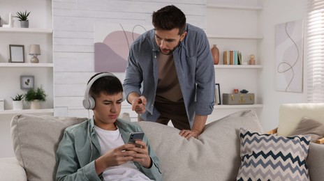 Photo of Father scolding his son while he listening music in headphones at home. Teenager problems