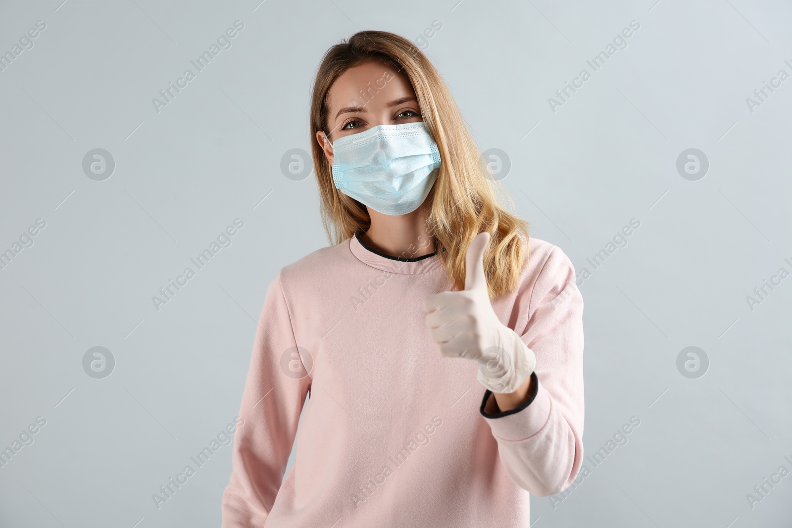 Photo of Young woman in medical gloves and protective mask showing thumb up on grey background