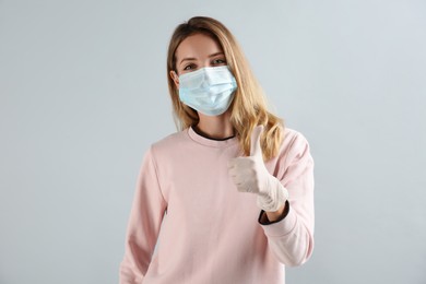 Photo of Young woman in medical gloves and protective mask showing thumb up on grey background