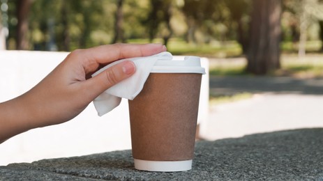 Photo of Woman cleaning lid of coffee cup with wet wipe outdoors, closeup. Protective measures