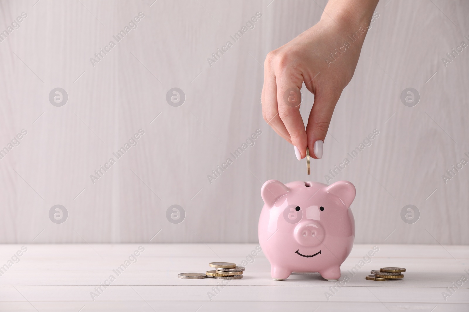 Photo of Woman putting coin into piggy bank at white wooden table, closeup with space for text