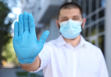 Photo of Man in protective face mask showing stop gesture outdoors, focus on hand. Prevent spreading of coronavirus