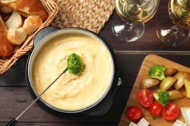 Dipping piece of broccoli into fondue pot with melted cheese at wooden table with wine and snacks, flat lay