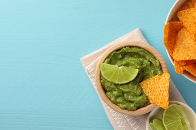 Bowl of delicious guacamole, lime and nachos chips on light blue wooden table, flat lay. Space for text