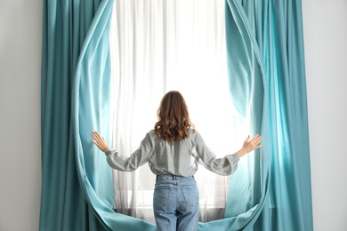 Woman opening window curtains at home in morning, back view