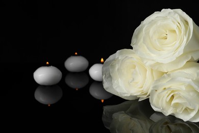 Photo of White roses and burning candles on black mirror surface in darkness, closeup with space for text. Funeral symbols