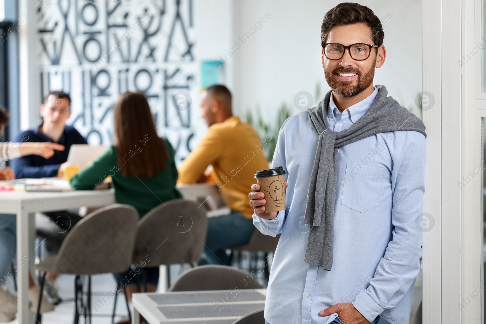 Photo of Team of employees working together in office. Happy man with paper cup of drink indoors, space for text