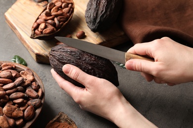 Photo of Woman cutting ripe cocoa pod over grey table with products, closeup