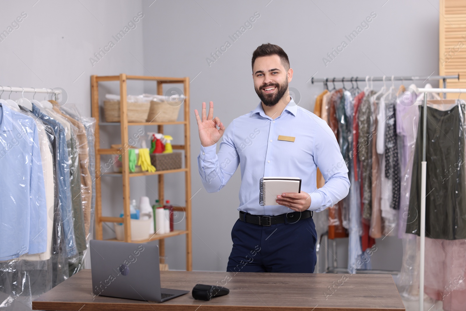 Photo of Dry-cleaning service. Happy worker with notebook showing ok gesture in workplace indoors