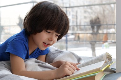 Photo of Cute little boy reading book near window at home