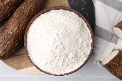 Bowl with cassava flour and roots on white table, top view