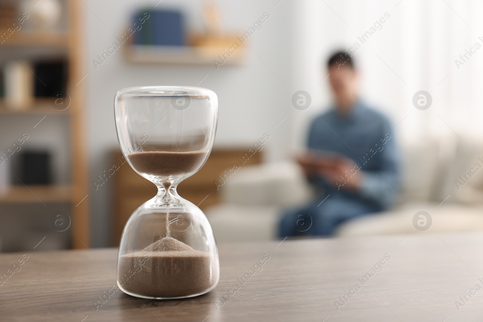 Photo of Hourglass with flowing sand on desk. Man reading book in room, selective focus