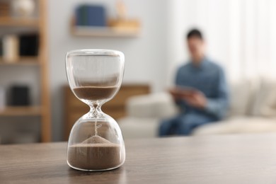 Photo of Hourglass with flowing sand on desk. Man reading book in room, selective focus