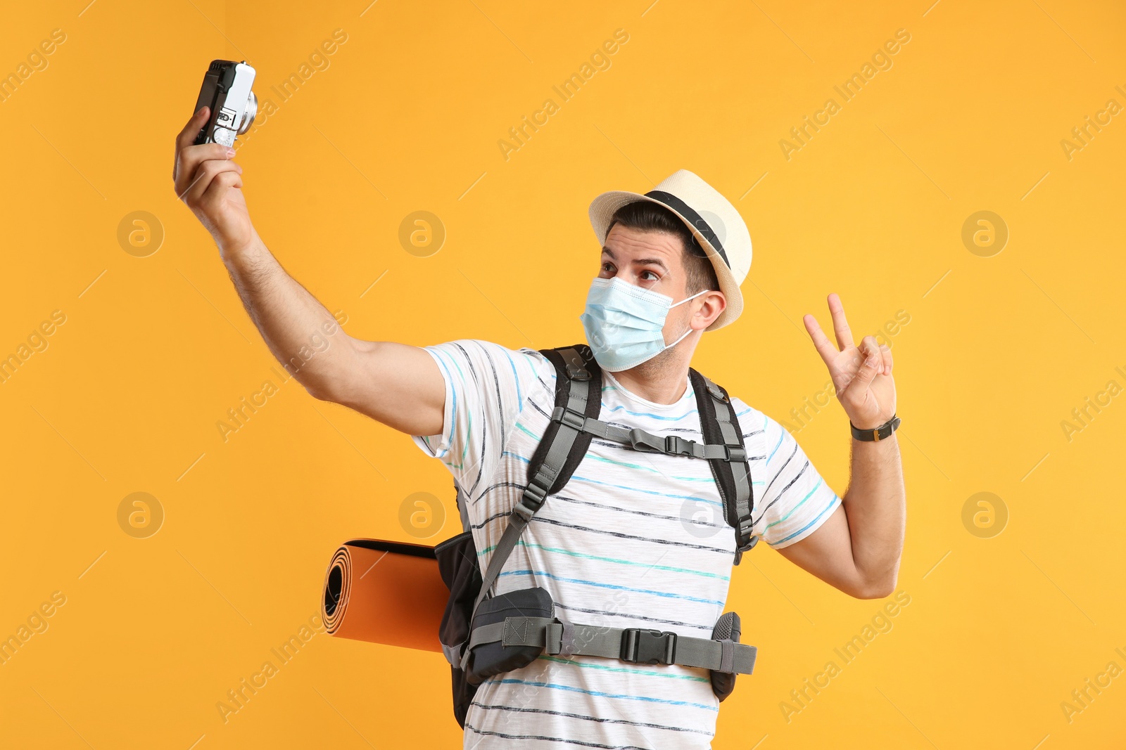 Photo of Male tourist in protective mask with travel backpack taking selfie on yellow background