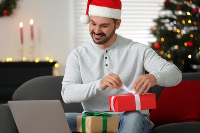 Celebrating Christmas online with exchanged by mail presents. Happy man in Santa hat opening gift box during video call on laptop at home
