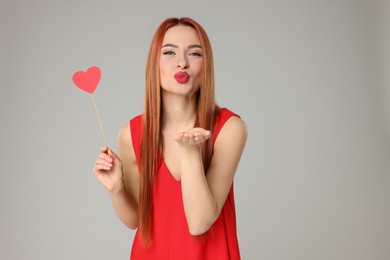 Photo of Young woman in red dress with paper heart blowing kiss on light grey background