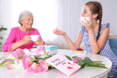 Photo of Preteen girl with her granny drinking tea at home, focus on flowers and card. Happy Mother's Day
