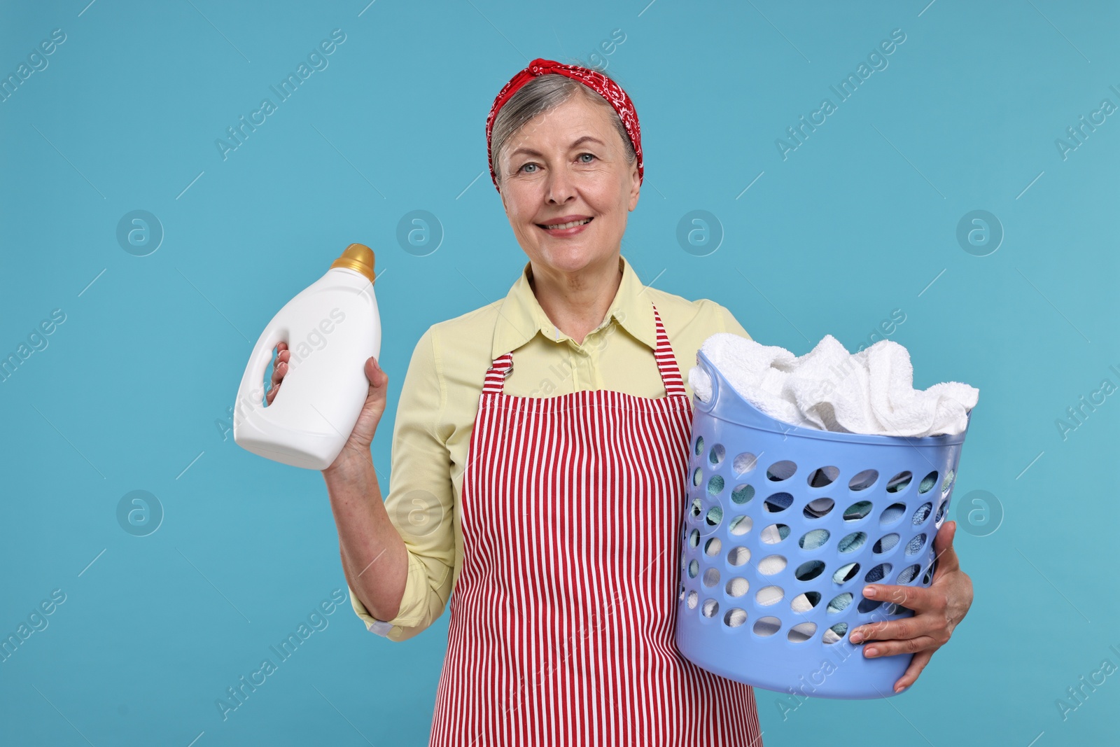 Photo of Happy housewife with detergent and basket full of laundry on light blue background