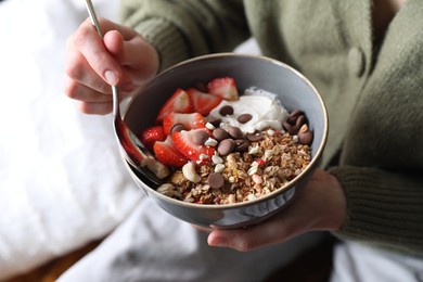 Woman eating tasty granola with chocolate chips, strawberries and yogurt indoors, closeup