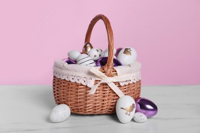 Photo of Wicker basket with festively decorated Easter eggs on white marble table against pink background
