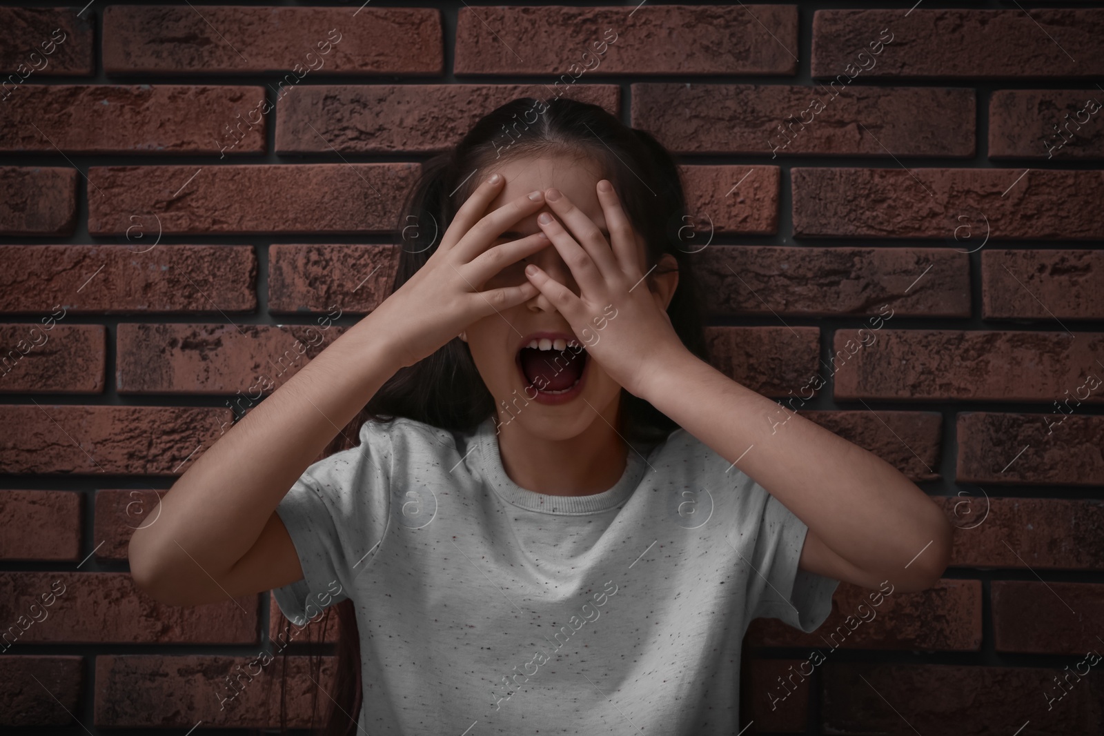 Photo of Scared little girl closing eyes with hands near brick wall. Child in danger